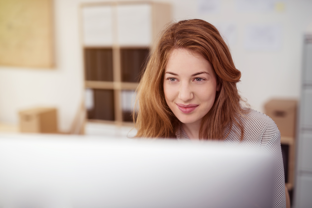 woman working on a desktop computer smiling as she leans forwards reading text on the screen, view over the monitor